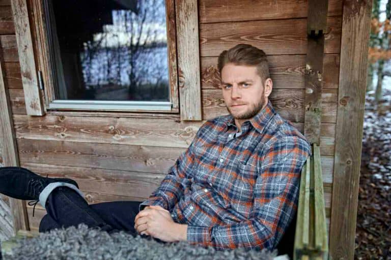 Mann trägt Karro Hemden Übergröße Herren sitzt entspannt vor rustikalem Holzhintergrund